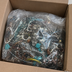 A bulk lot of assorted jewelry, including necklaces, bracelets, and earrings with beads, pearls, and turquoise accents, packed inside a clear plastic bag and sitting in an open cardboard box.