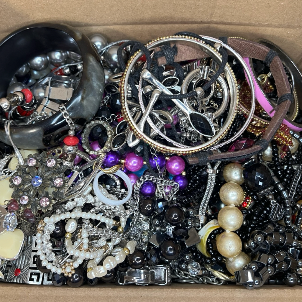 A bulk lot of assorted vintage and thrift jewelry pieces piled in a cardboard box, including silver and gold-toned chains, beaded necklaces, pearl strands, bracelets, and earrings.