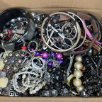 A bulk lot of assorted vintage and thrift jewelry pieces piled in a cardboard box, including silver and gold-toned chains, beaded necklaces, pearl strands, bracelets, and earrings.