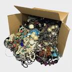 A large assortment of mixed jewelry, including pearl strands, beaded necklaces, bracelets, and earrings, piled out of an open brown cardboard box on a white background.