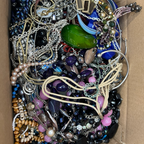 A cardboard box filled with assorted jewelry, including beaded necklaces, chains, bracelets, rings, and earrings in black, blue, pink, purple, green, silver, and gold tones.