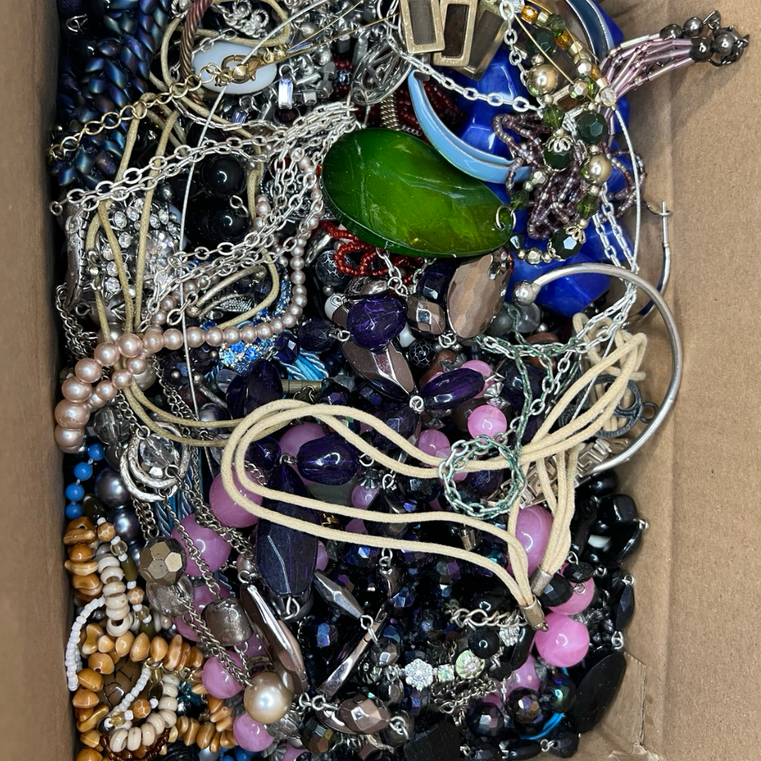 A cardboard box filled with assorted jewelry, including beaded necklaces, chains, bracelets, rings, and earrings in black, blue, pink, purple, green, silver, and gold tones.
